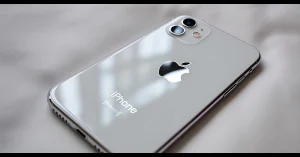 صورة مقربة لهاتف آيفون فضي اللون على قماش ناعم فاتح اللون. شعار Apple بارز، مع كاميرتين مرئيتين. تصميم أنيق وعصري.